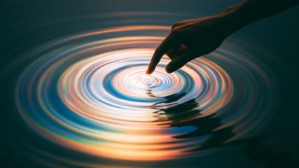 A silhouetted hand touches still water, creating colorful concentric ripples that glow against a dark teal background.