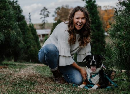 A photograph of Lauren crouching next to her dog among pine trees