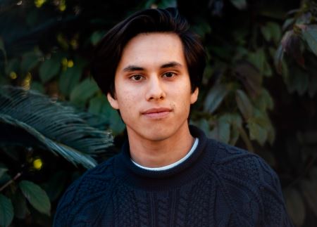 A close-up photo of Krzysztof wearing a blue sweater on a background of green foliage