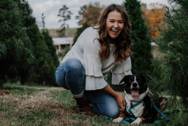 A photograph of Lauren crouching next to her dog among pine trees