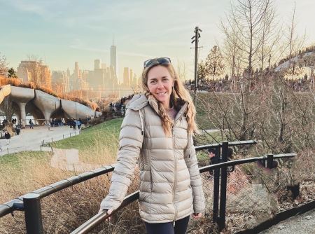 A photo of Amy wearing a cream-colored jacket outdoors in a park, with a city skyline in the background