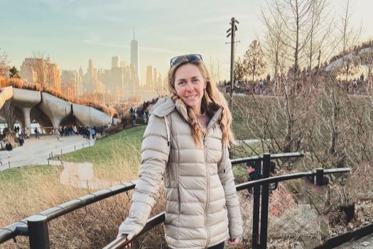 A photo of Amy wearing a cream-colored jacket outdoors in a park, with a city skyline in the background