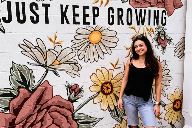 Sasha standing in front of a decorative wall with the text "Just Keep Growing"