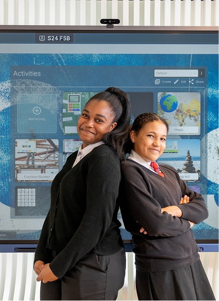 Two confident female students in school uniforms standing back-to-back in front of interactive digital display
