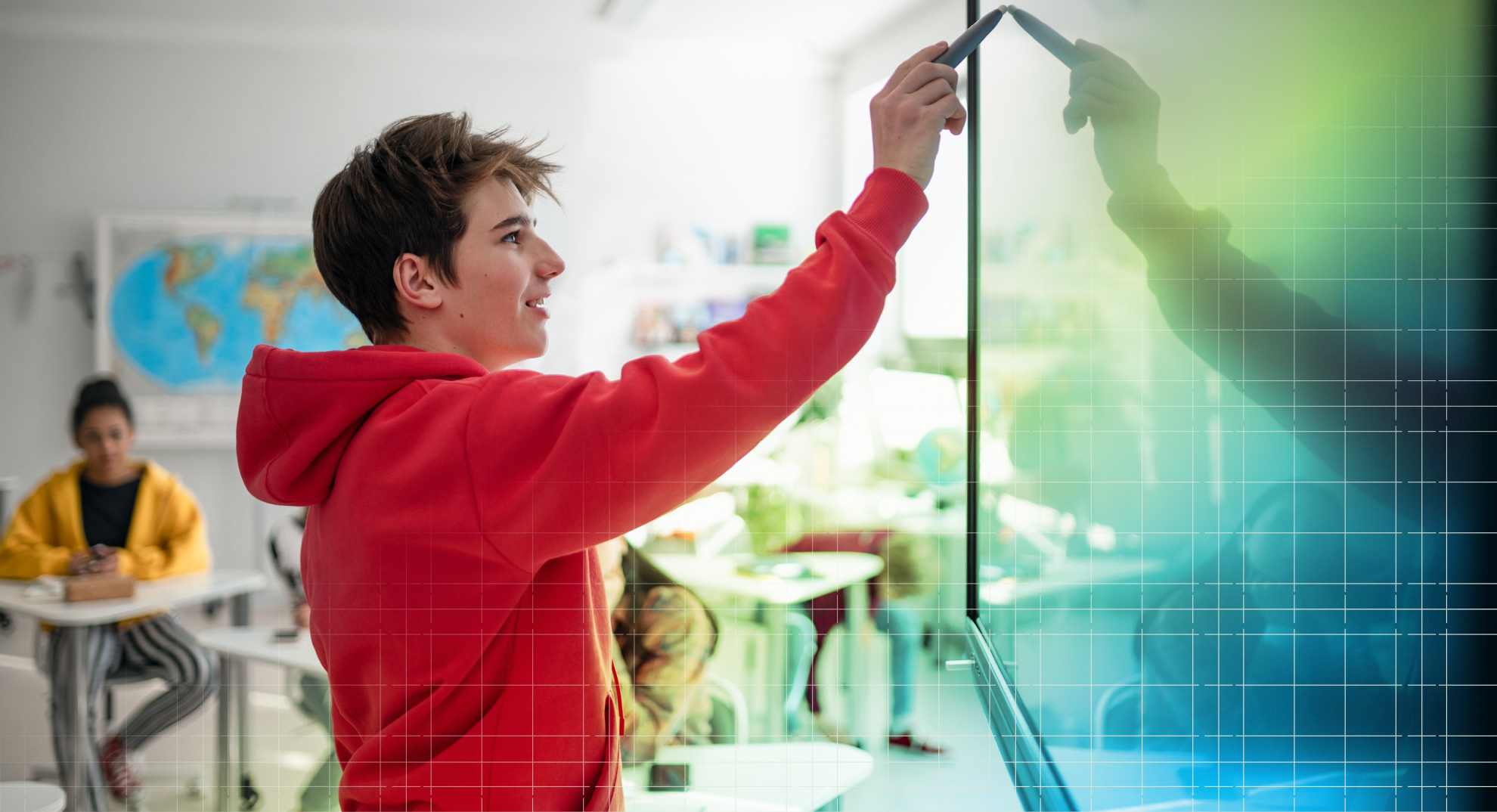 Smiling student in red hoodie writing on interactive digital touchscreen display in modern classroom with world map and classmate in background