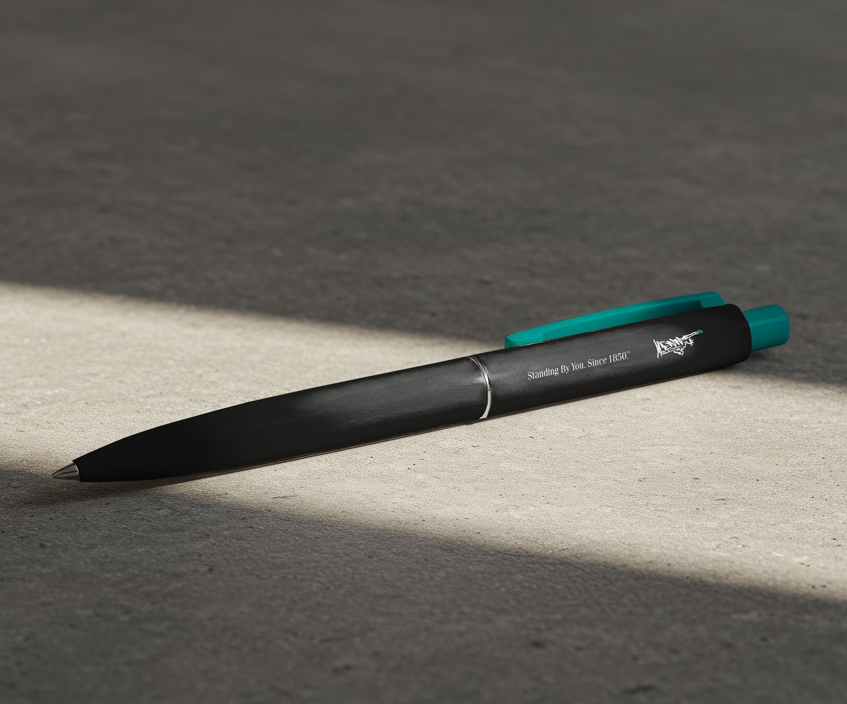 Black ManhattanLife pen with teal clip lying in a beam of light on concrete surface.