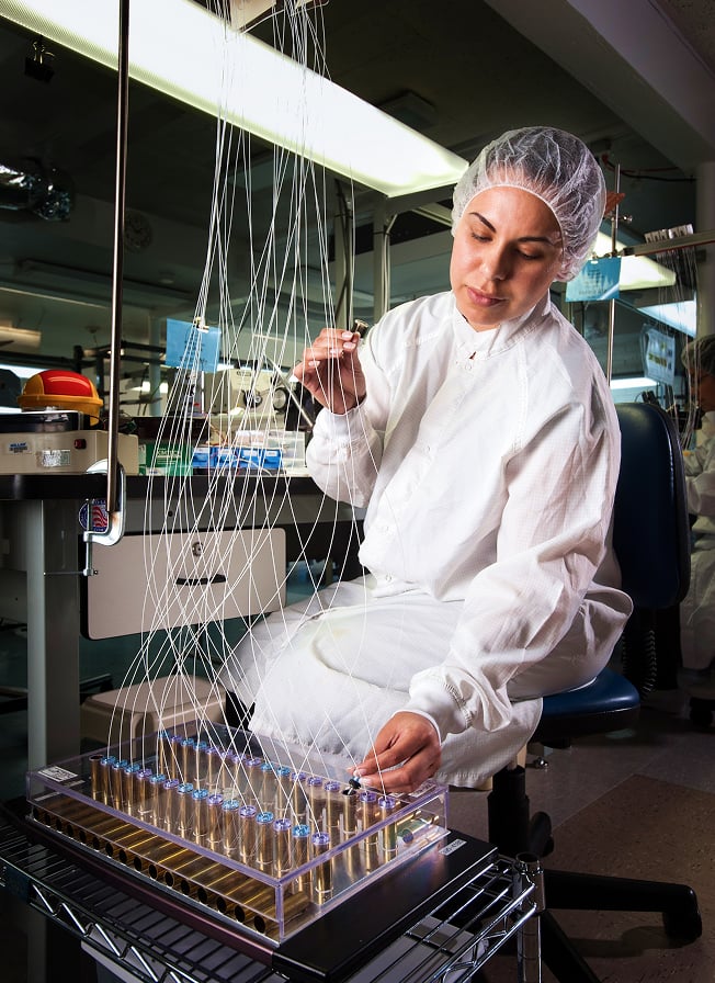 Lab technician in cleanroom suit inspecting multiple thin sensor wires connected to test tubes in a medical device lab.
