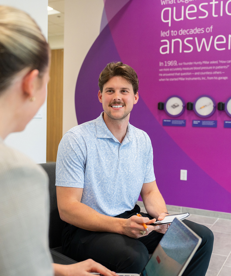 Man and colleague chatting in front of a purple Millar history wall display featuring text and framed medical device artifacts.