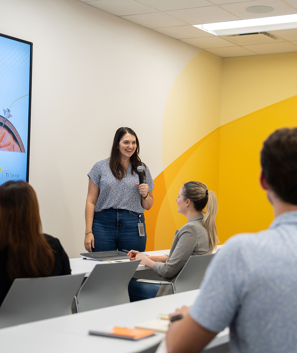 Woman leading a training session with microphone in a Millar classroom, speaking to colleagues in front of a screen and yellow branded wall.