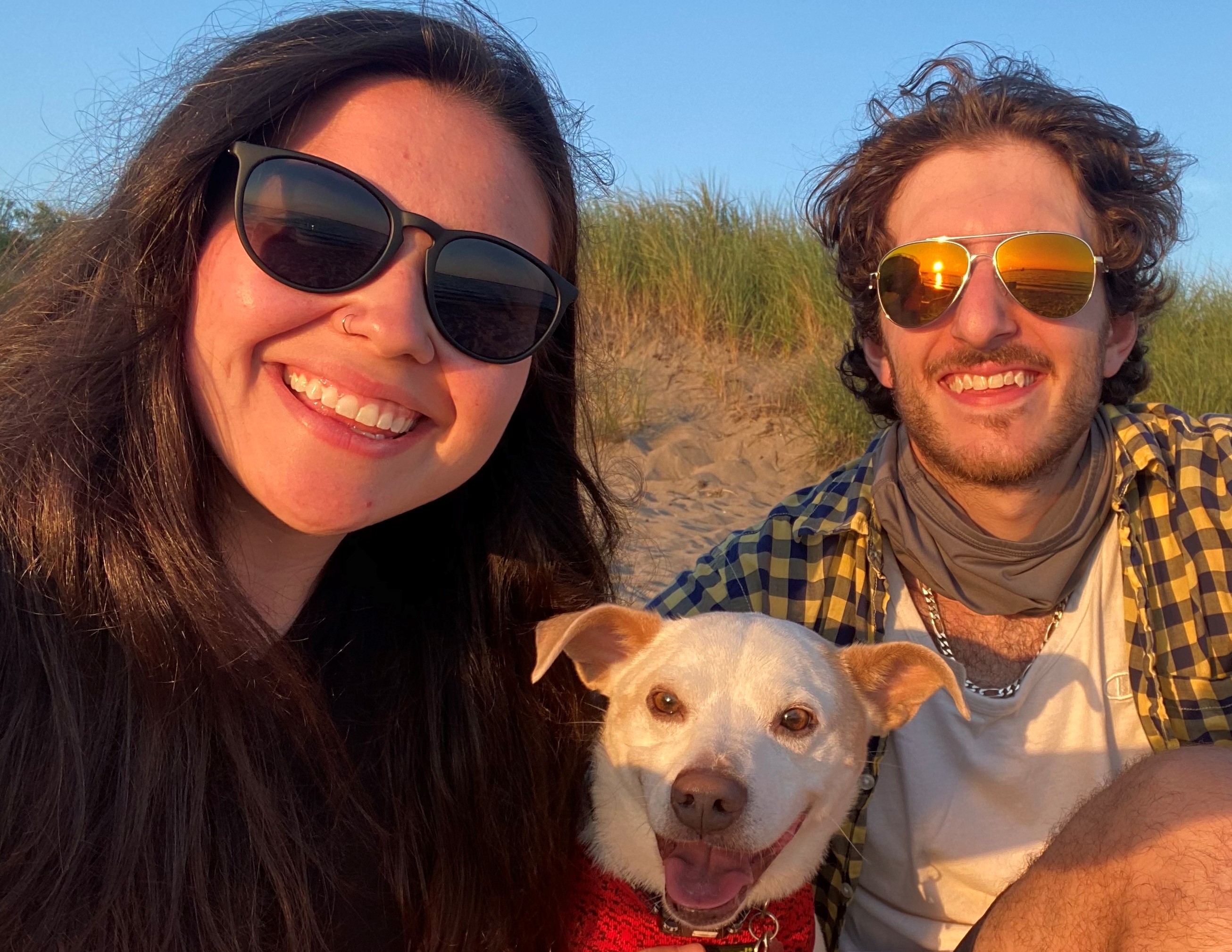 Anthony sitting with his fiance and dog on a sandy hill, blue sky in the background.