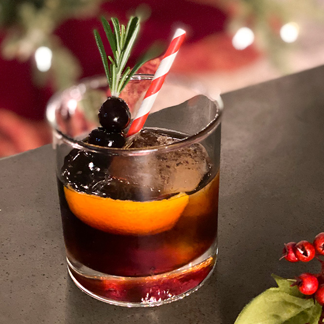 A dark amber drink garnished with rosemary and cherries, served in an old fashioned style glass with a candy cane patterned straw.