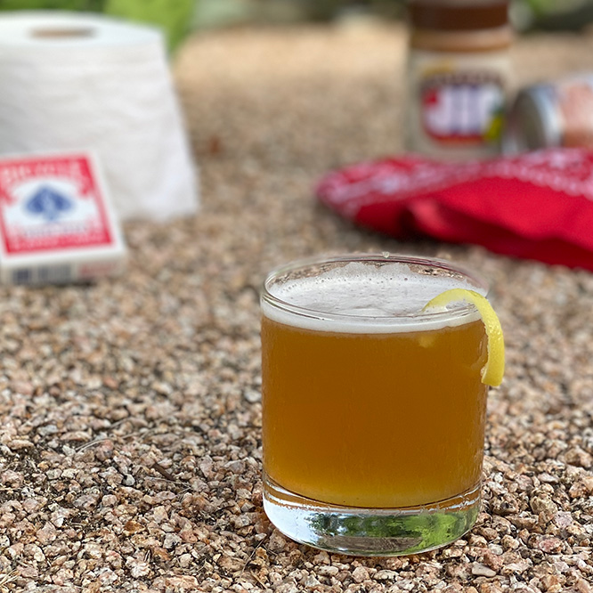 A golden brown drink with a thin layer of froth on top in an old-fashioned glass, with a lemon twist on the rim.