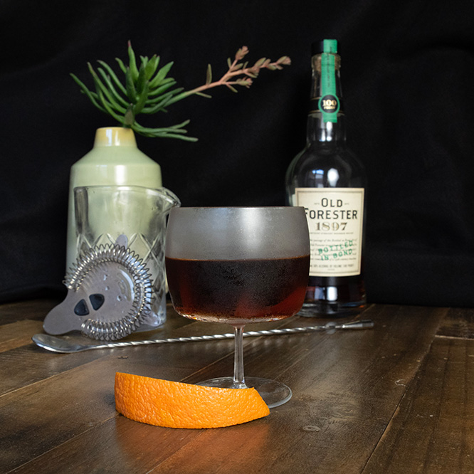 A dark crimson colored drink in a goblet style glass, with an orange peel alongside the glass.