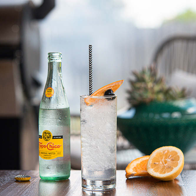 A clear drink in a collins glass with a striped straw, garnished with a cherry and a lemon wedge.